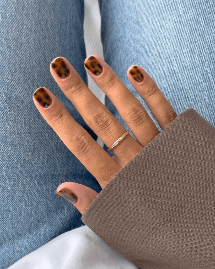 Close-up of a hand with brown and black spotted nail art, wearing a silver ring, against a background of denim and fabric.