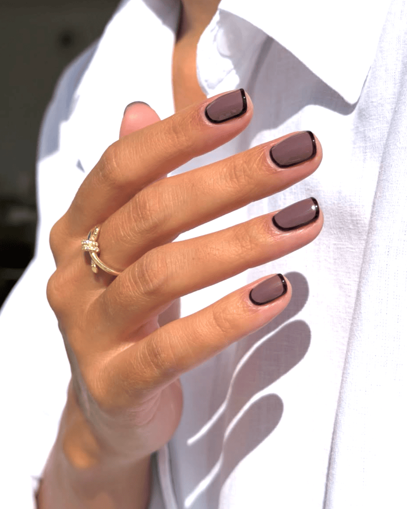 A close-up of a hand with elegantly manicured nails featuring a dark mauve color and black tips, adorned with a gold ring, against a light-colored shirt.