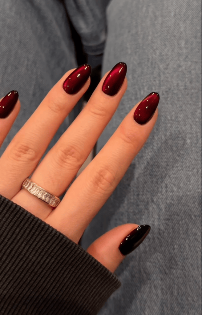 Close-up of a hand with elegantly manicured nails featuring a gradient of deep red to black. A silver ring is worn on one finger, and the hand is partially covered by a dark sweater, with a soft focus on denim fabric in the background.
