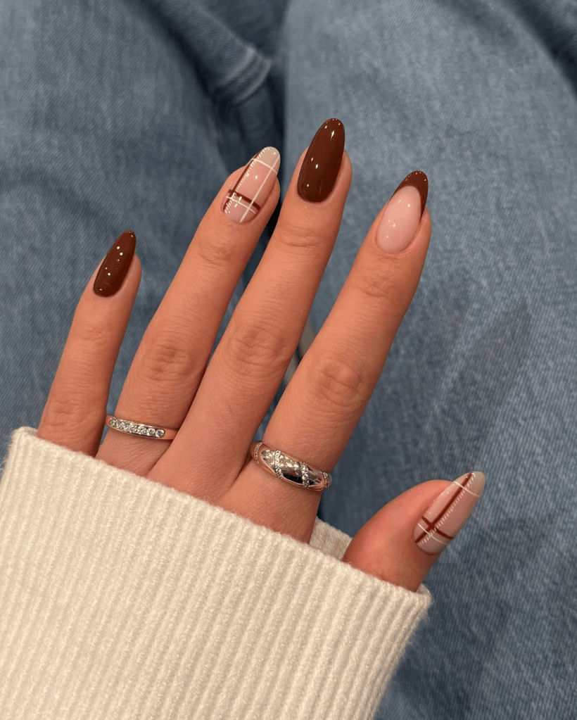 A close-up of a hand showing stylishly manicured nails, featuring a mix of dark brown, nude, and patterned designs. The hand is resting on a soft, cream-colored sweater, with visible silver rings on two fingers.