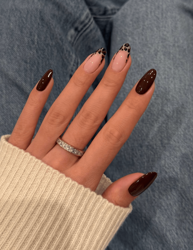 A hand showing manicured nails with a mix of dark brown and nude polish, featuring decorative black accents, while wearing a silver ring. The hand is partially covered by a cream-colored sweater, against a backdrop of denim fabric.