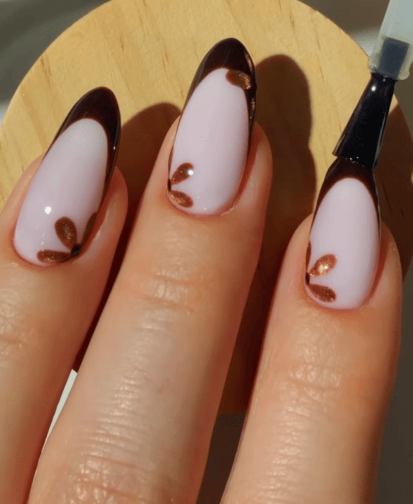 Close-up of manicured nails featuring a light pink base with dark brown tips and small bronze accents.