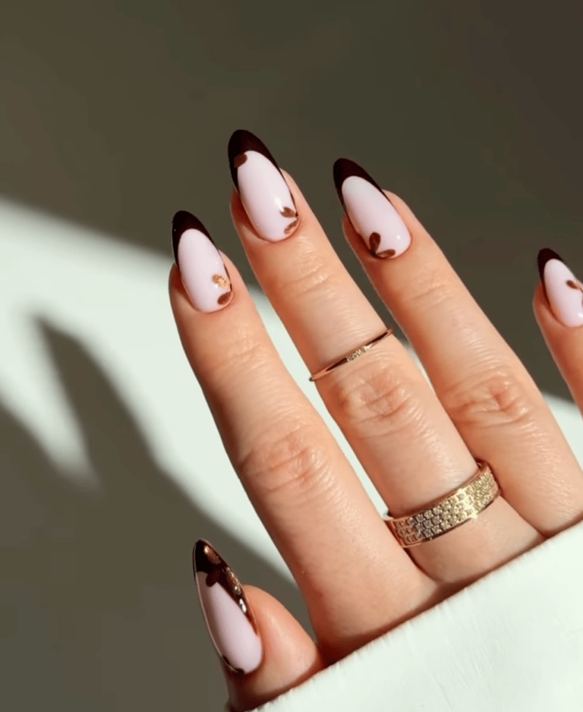 Close-up of a hand with elegantly manicured nails featuring pink and dark brown tips. The fingers are adorned with delicate rings, and the hand is positioned against a light background.