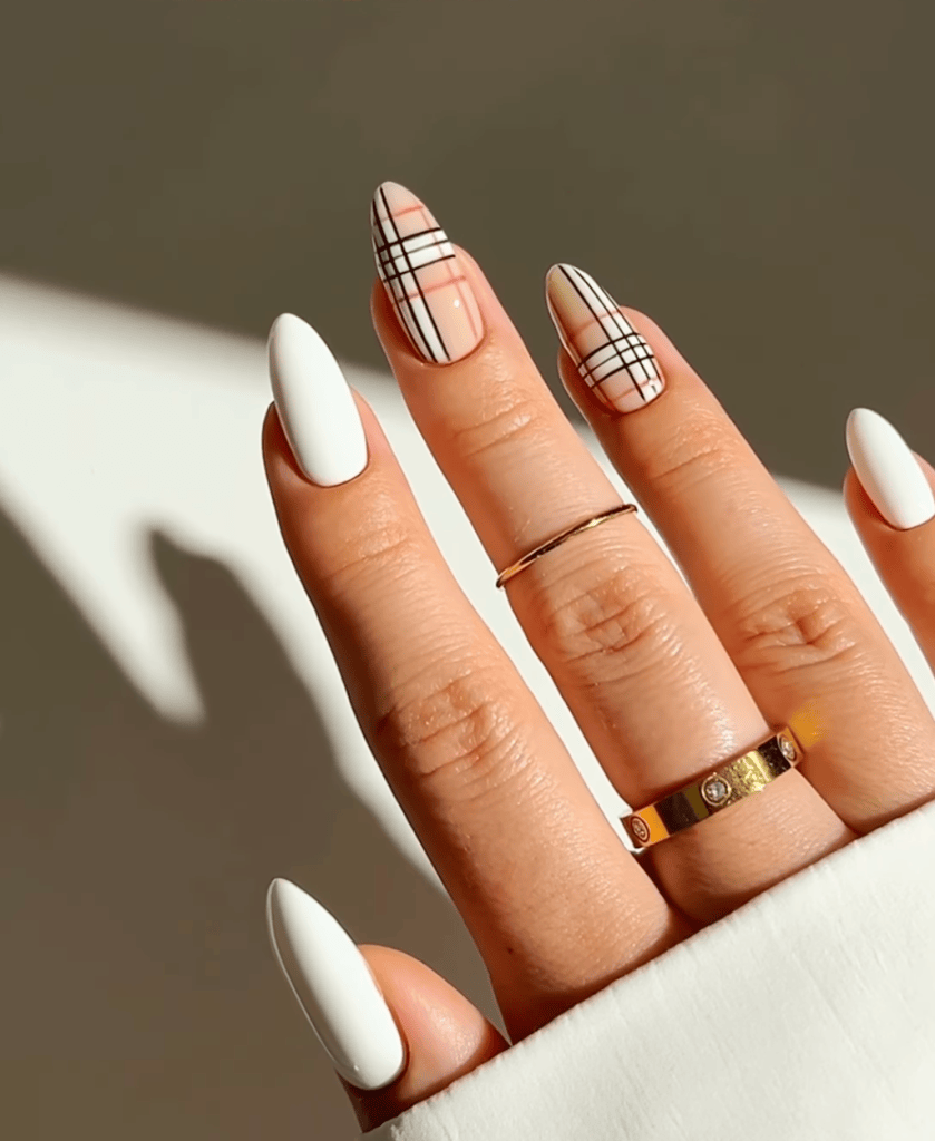 Close-up of a hand with long, pointed white and patterned nails. The nails feature a plaid design on one finger. The hand is adorned with several rings.