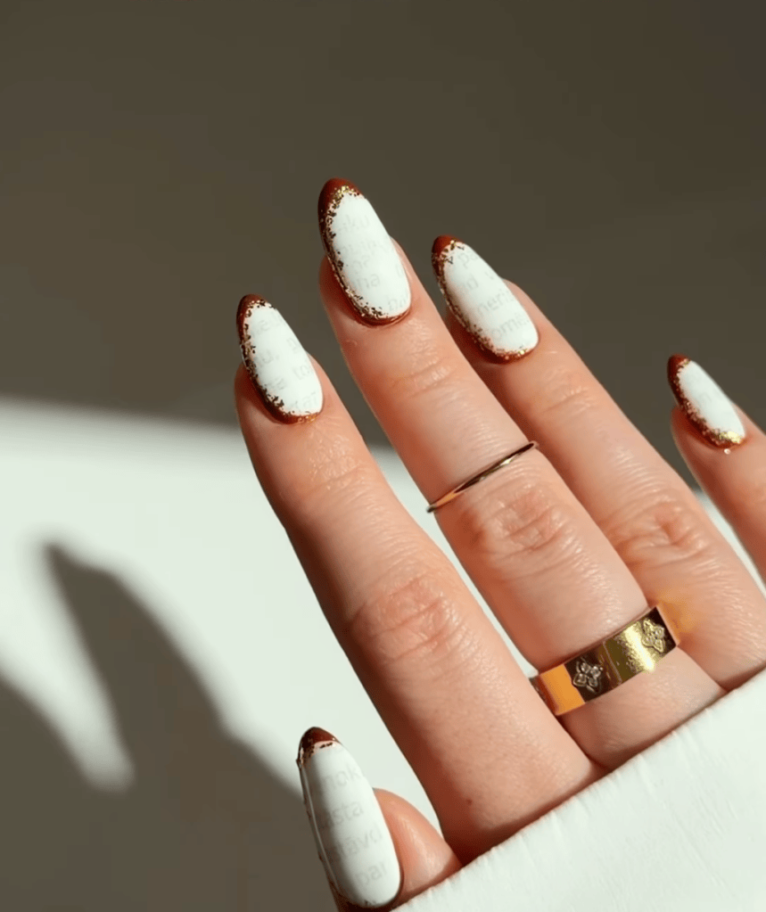 Close-up of manicured nails with white polish featuring text designs, gold foil tips, and worn gold rings on fingers.