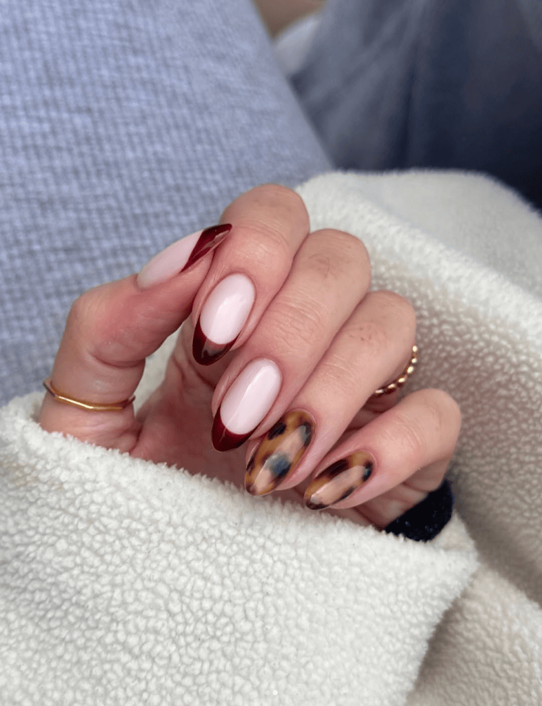 Close-up of a hand displaying stylish nails with a mix of dark red tips, pale pink, and tortoiseshell designs while resting on a soft beige fabric.