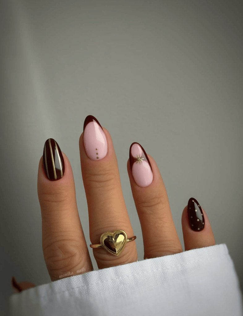 Close-up of a hand with elegantly manicured nails featuring a mix of dark brown and pink colors, including nail art designs such as stripes, dots, and a star. A gold heart-shaped ring is worn on the ring finger, with a white fabric visible at the bottom.