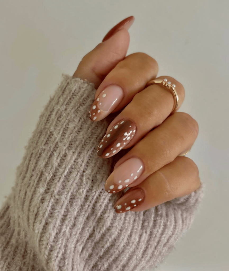 Close-up of a hand with stylish brown and nude ombre nails featuring white polka dots, wearing a cozy beige sweater and a gold ring.