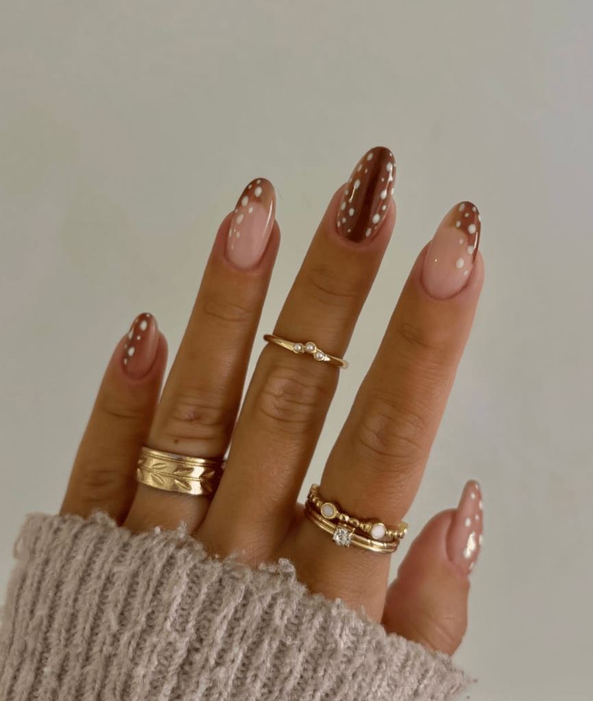 Close-up of a hand with stylishly manicured nails featuring brown and nude colors, adorned with white polka dots. The hand is accessorized with multiple gold rings, and the person is wearing a cozy gray sweater.