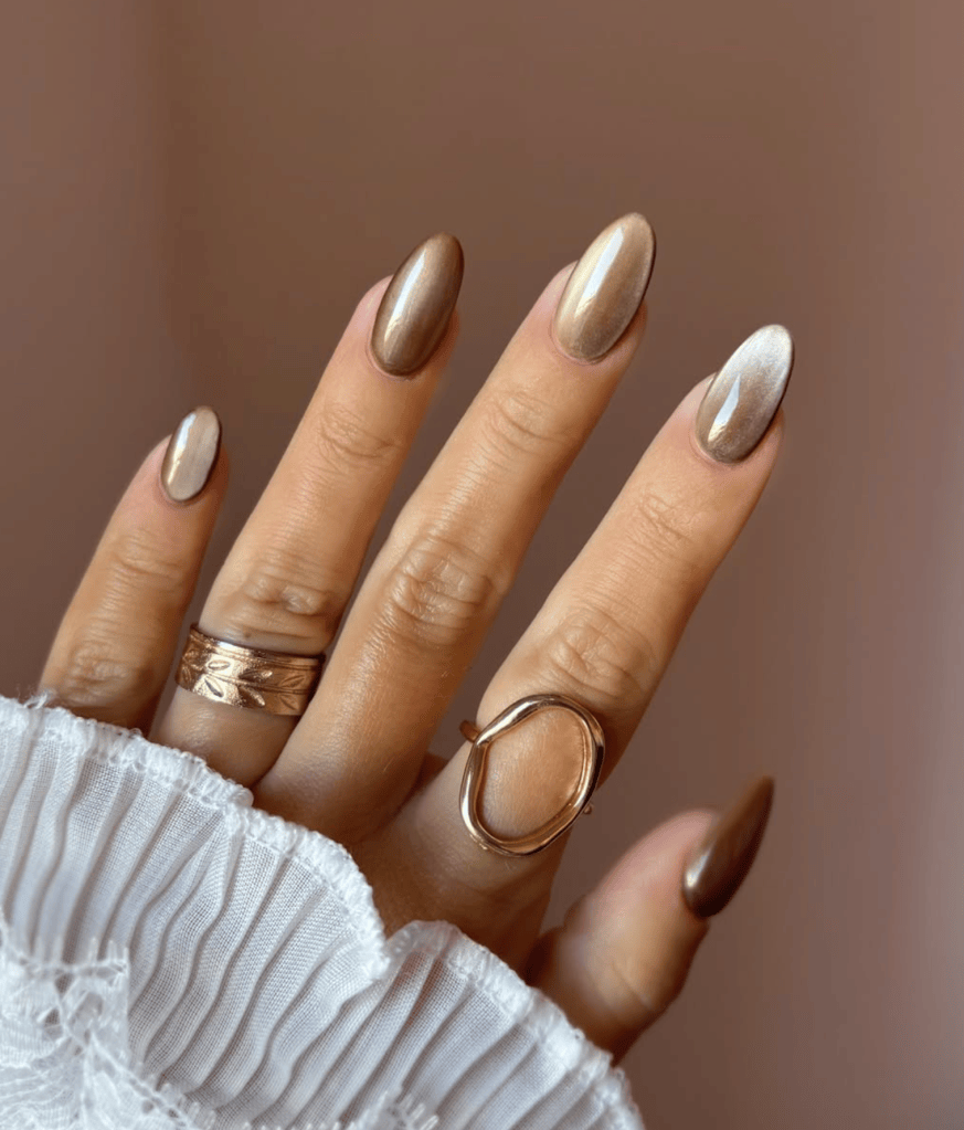 Close-up of a hand with stylish almond-shaped nails painted in metallic brown and a shiny accent nail. The hand is adorned with multiple rings and is partially covered by a white ruffled fabric.