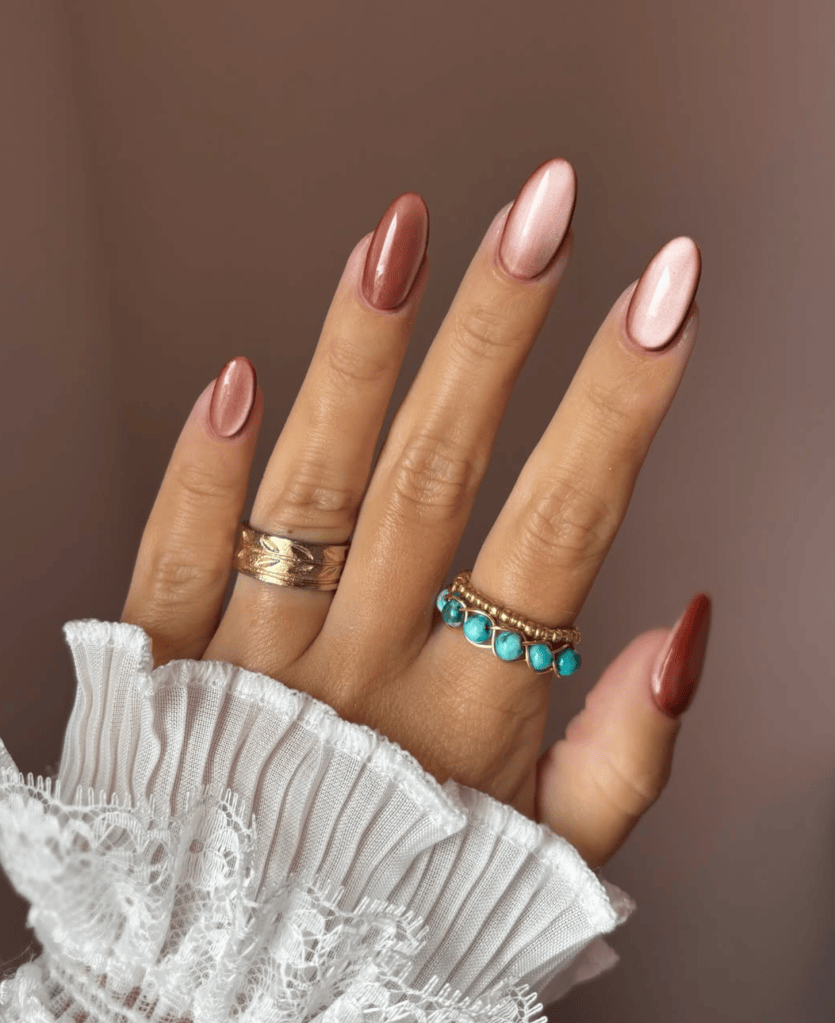 A close-up of a hand with elongated, manicured nails in shades of rose gold and pink, wearing three rings: a gold textured ring, a gold ring with turquoise stones, and a decorative gold band, all paired with a white ruffled blouse.