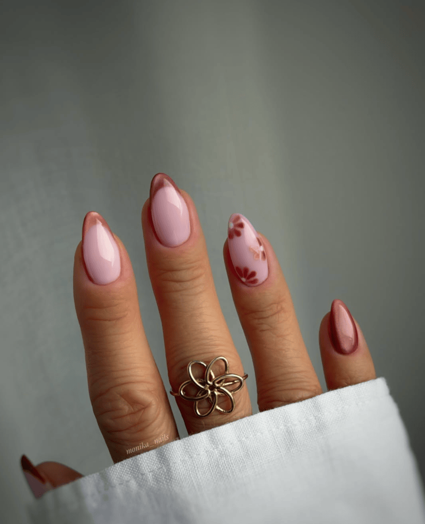 A close-up of a hand with elegantly manicured nails in shades of pink, featuring a floral design on one nail and a gold ring with a floral pattern.