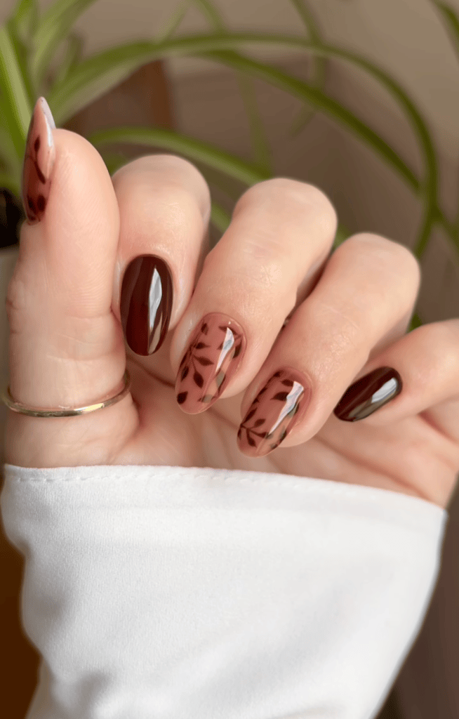 Close-up of a hand displaying stylish nail art featuring dark brown polished nails and two accent nails with leaf designs in a lighter brown.