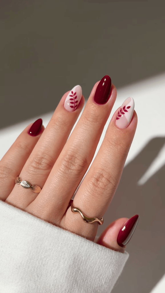 Close-up of a hand with manicured nails featuring burgundy and white polish, decorated with leaf patterns, and wearing two rings.