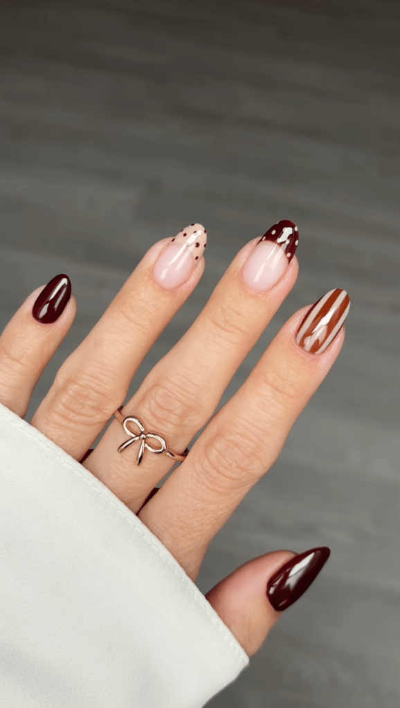 A close-up view of a hand displaying beautifully manicured nails in various designs, including dark red, pink, and chocolate brown tones, along with a ring shaped like a bow.