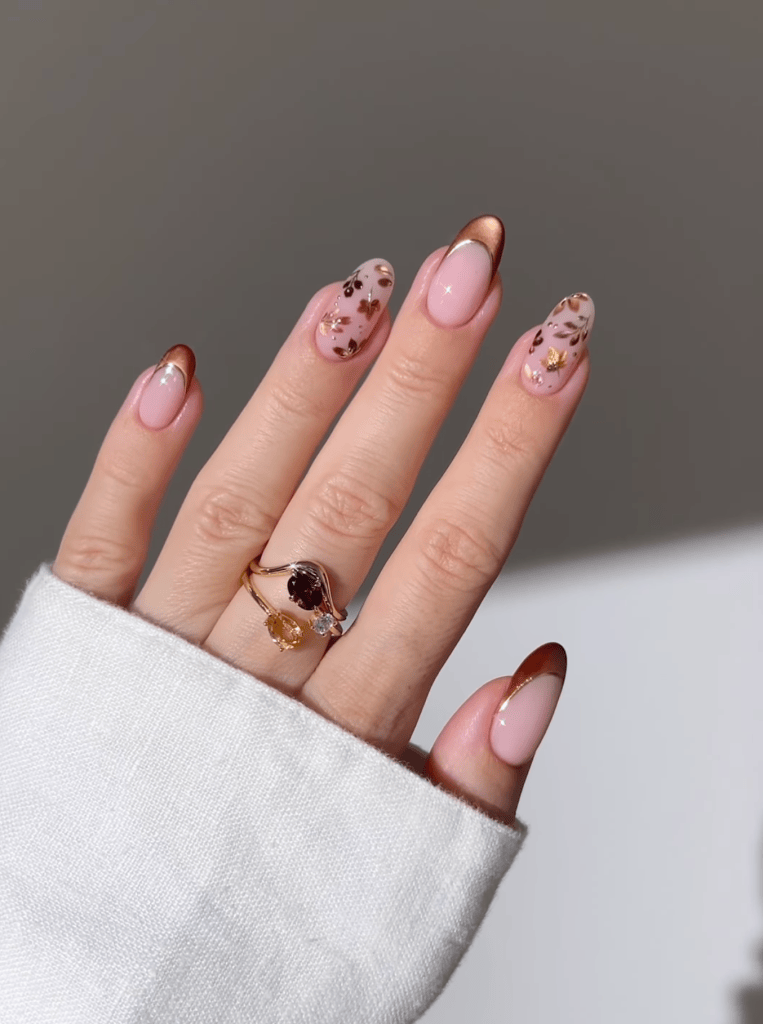 A close-up of a hand with well-manicured nails featuring a pink base, brown tips, and decorative floral designs. The hand is adorned with a stylish ring showcasing multiple gemstones, and the wrist is partially covered by a white sweater.