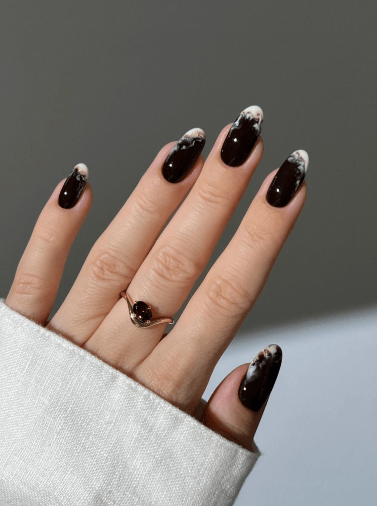 A hand showing long, dark brown nails with white tip designs and a silver ring on the ring finger, partially covered by a light sweater.
