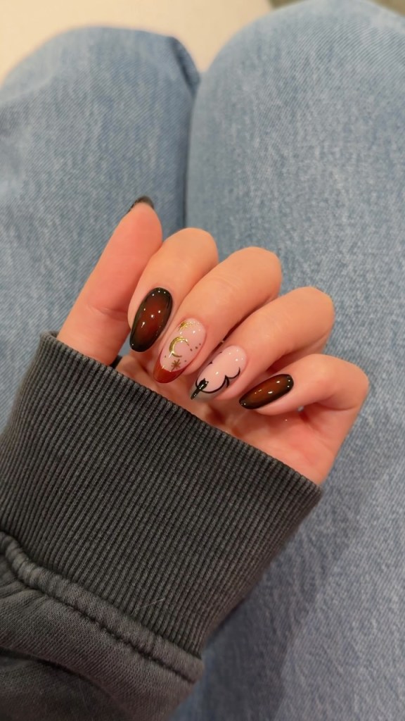 A close-up of a hand showcasing beautifully manicured nails in various designs, including dark ombre, soft pink with glitter, and intricate patterns. The hand is resting on a grey sweater, with denim jeans visible in the background.