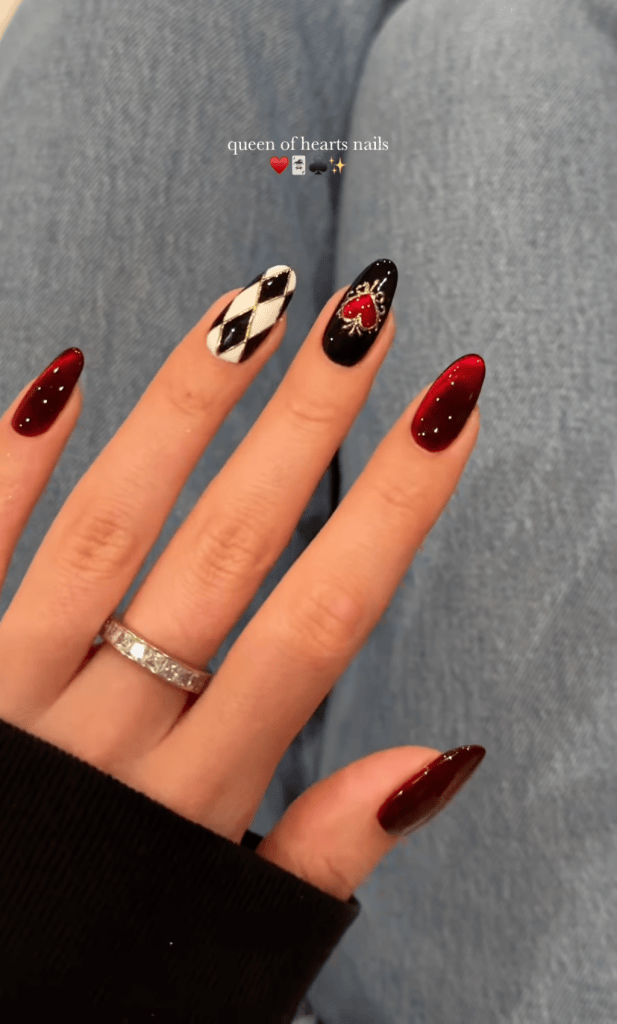 A hand with stylish, long nails featuring a 'Queen of Hearts' theme. The nails are painted in red and black, with one nail displaying a diamond pattern and another featuring a heart design adorned with rhinestones. A silver ring is visible on the finger, with denim jeans in the background.