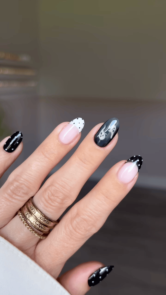 A close-up view of a hand with manicured nails featuring a mix of colors and designs, including black, pink, and white with polka dots and a bunny graphic on one nail. The hand is adorned with several gold rings.
