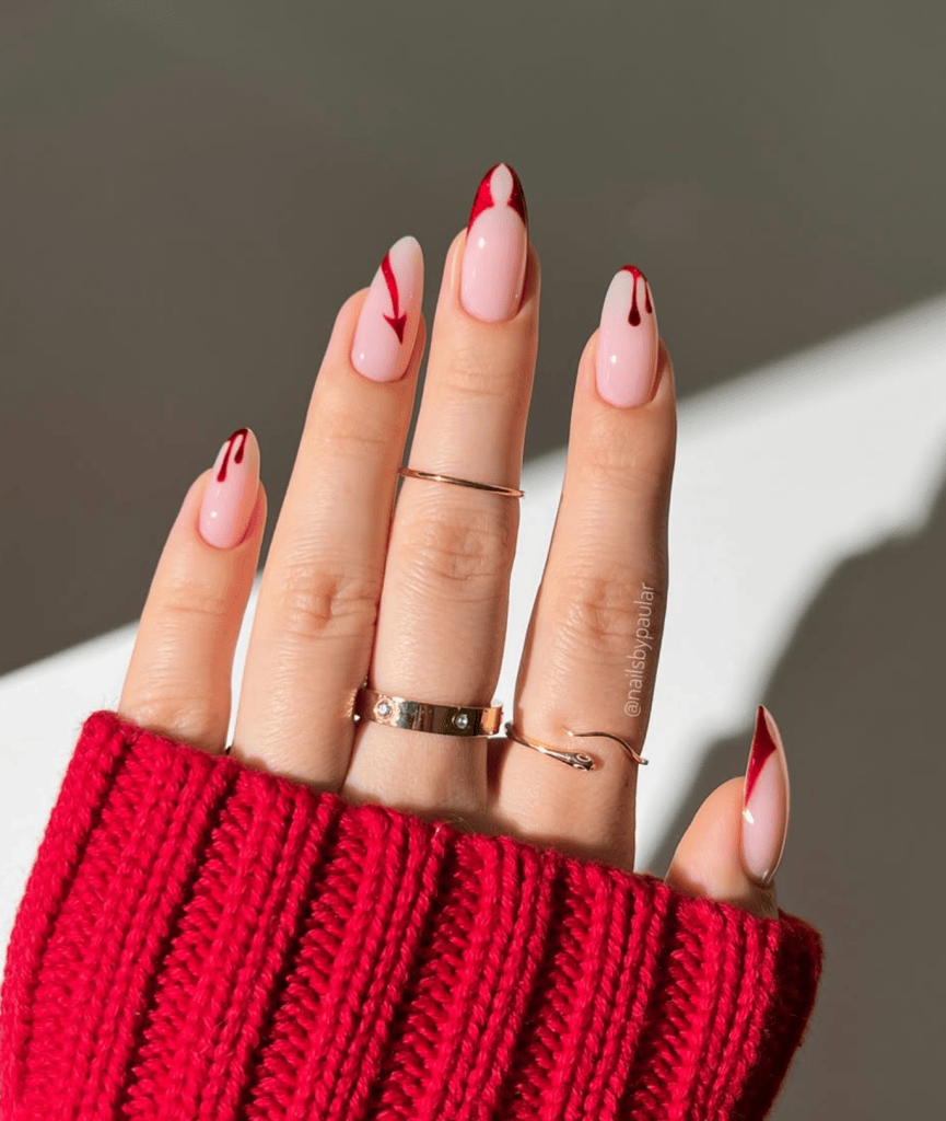 A close-up of a hand with elegantly manicured nails featuring a pink base and striking red tips, adorned with decorative red lines. The hand is wearing multiple rings and resting on a red knitted sweater.