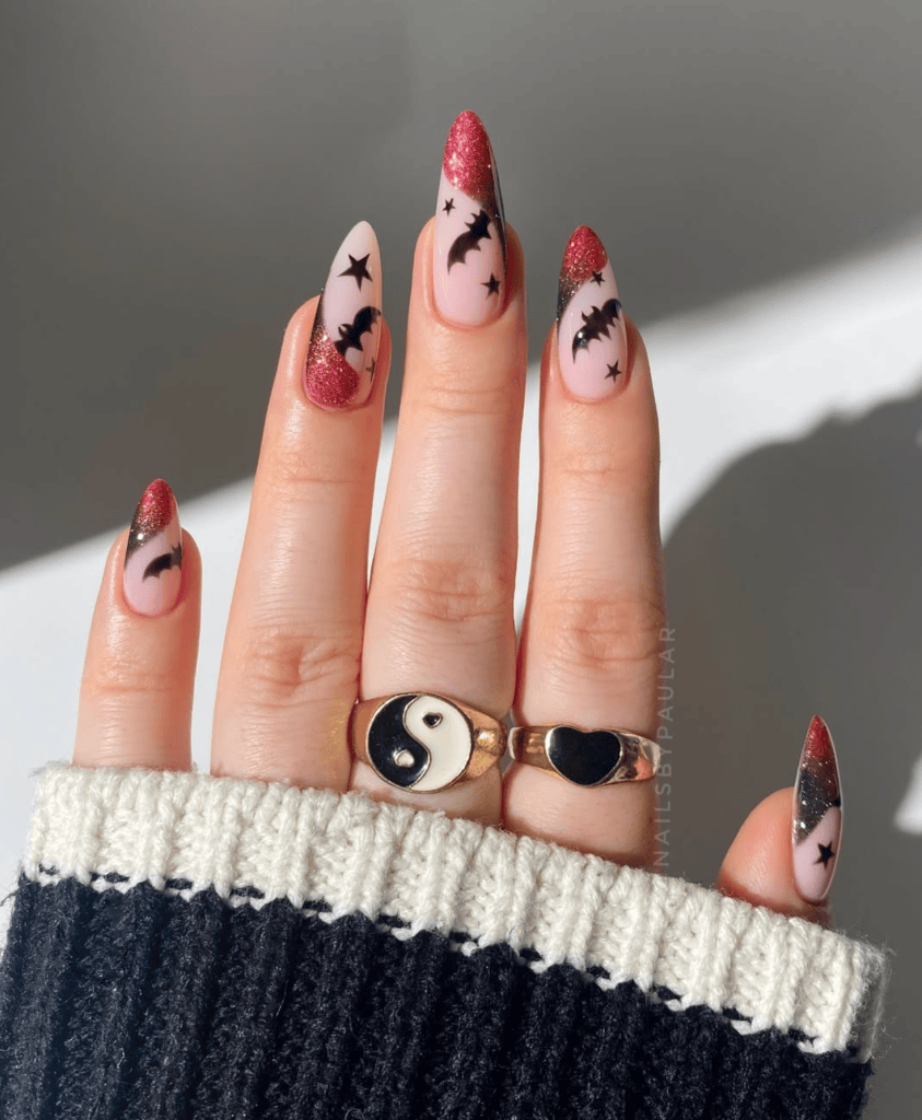 close-up of elegantly manicured nails with a Halloween-themed design featuring black bats and stars, showcasing a gradient of red glitter at the tips, paired with gold rings featuring a yin yang and a heart symbol