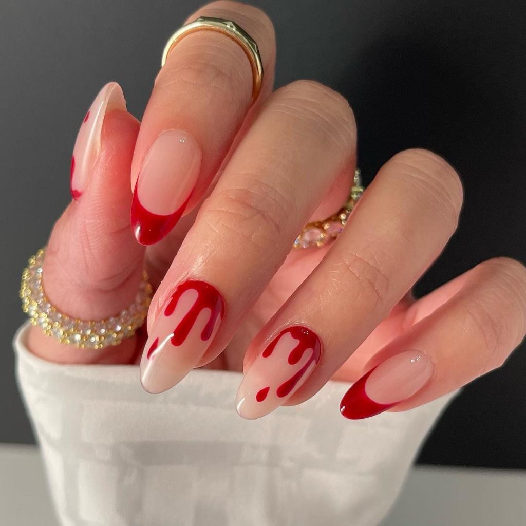 A close-up of a hand with long, almond-shaped nails featuring a gradient design in nude and red, with red dripping nail art at the tips. The fingers are adorned with gold rings and a glittery bracelet.