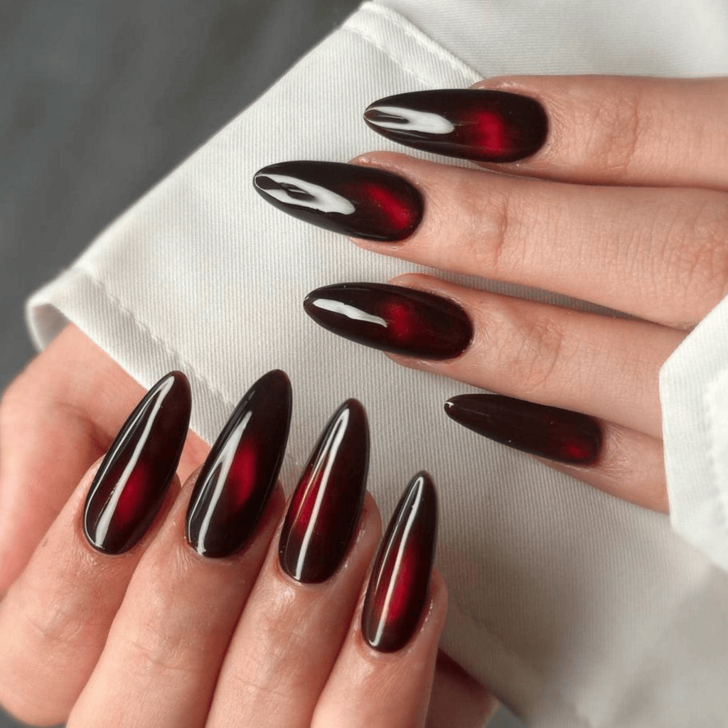 Close-up of manicured hands featuring long, pointed nails with a deep red and black gradient polish, showcasing a glossy finish.