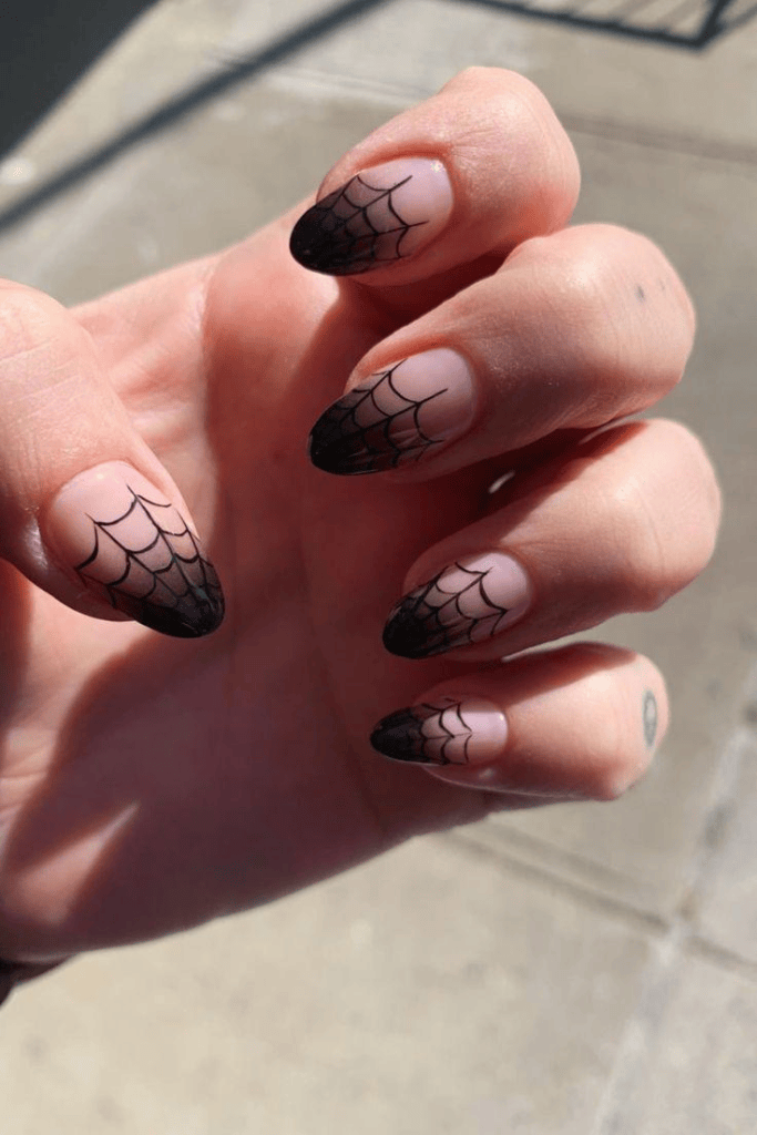 A close-up of a hand with manicured nails featuring a gradient design. The tips are painted black and have a spider web pattern, while the base is a natural nude shade.