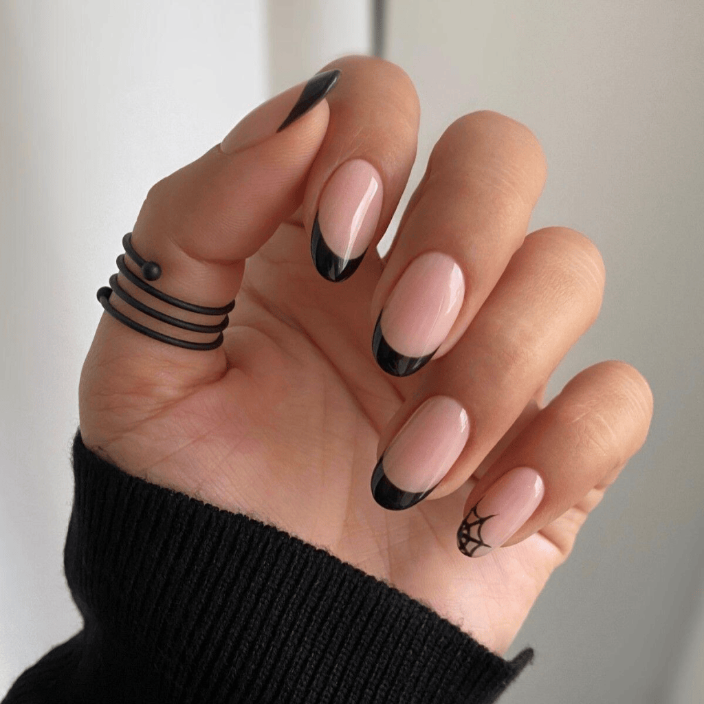 A hand displaying neatly manicured nails with a nude base and black French tips, featuring a spider web design on one nail, paired with black wire rings and wearing a black sweater.