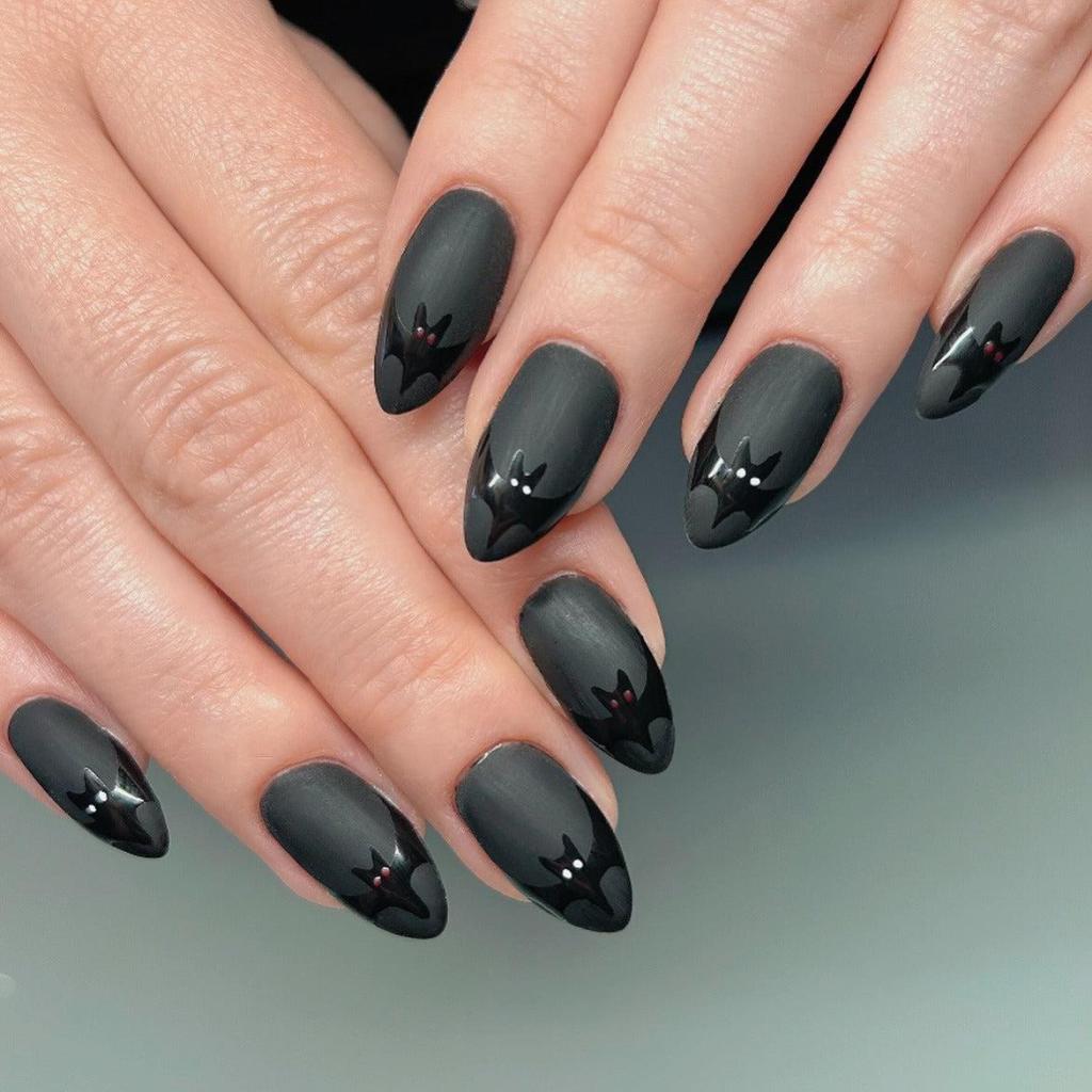 Close-up of manicured hands displaying black matte nail art with bat designs and accents in red and white.