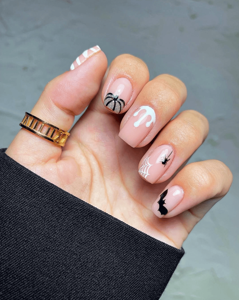 A close-up view of a hand with stylish Halloween-themed nails featuring a spider, spider web, bat, and dripping white design on a nude base. The person is wearing a gold and black ring.