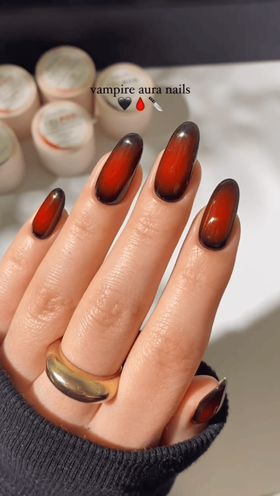 Close-up of a hand with long vampiric aura nails in a gradient of black to deep red, wearing a gold ring, against a blurred background of nail products.