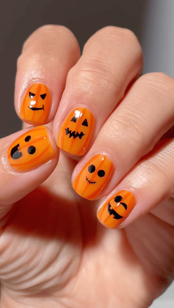 Close-up of fingernails painted orange with various Halloween-themed designs, including pumpkins and faces.