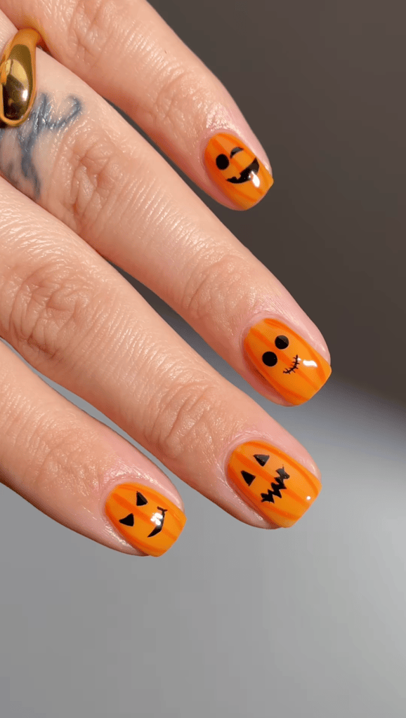 Close-up of orange Halloween-themed nail art featuring various pumpkin faces, including a smiling pumpkin, a jack-o'-lantern, and other playful designs on short nails.