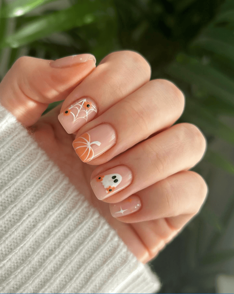 A hand showing a manicure with fall-themed nail art, including a pumpkin, spider web, ghost, and stars, set against a light background with greenery.