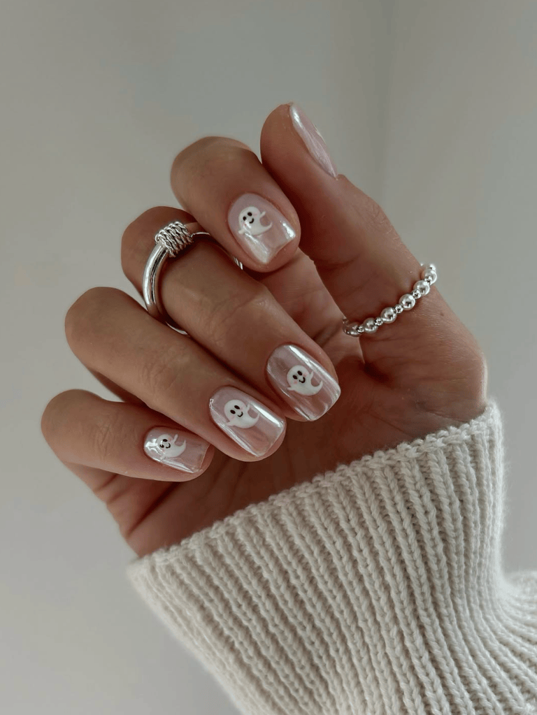 A hand showing off manicured nails with a light pink base and white ghost designs. The fingers are adorned with silver rings, and the background features a soft, neutral tone.